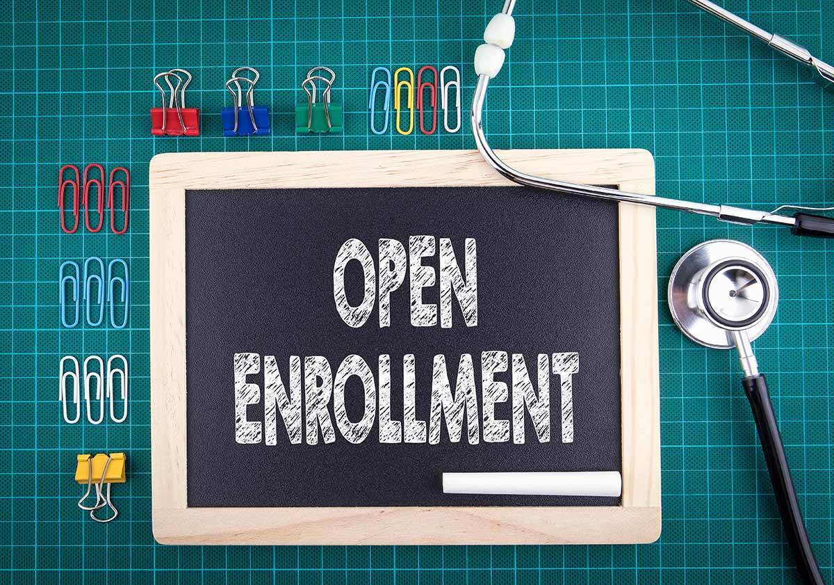 Slate writing board with chalk that says "open enrollment" with a doctor's stethascope and other reminder icons.